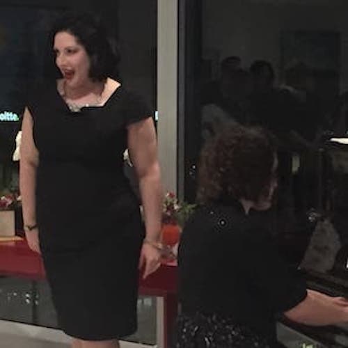 Leah wearing a black formal dress, standing next to a piano and singing while someone else plays the piano.