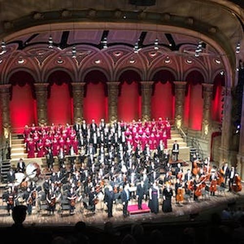 An aerial shot of an orchestra with a choir behind it and four people standing in the front of the stage and singing.